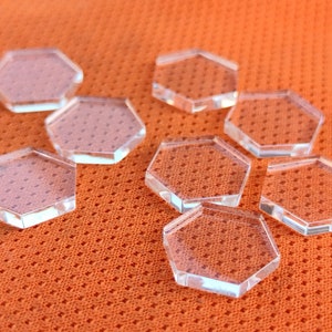 May include: Eight clear hexagonal acrylic bases, ideal for displaying miniature figures or other small objects. The bases are arranged in a staggered pattern on a textured orange surface.