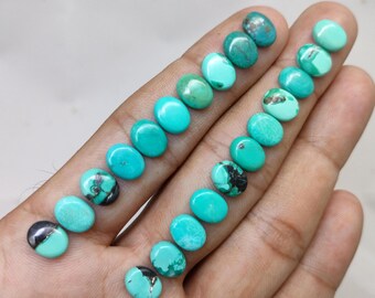 May include: A collection of oval-shaped turquoise gemstones in varying shades of blue and green. Some stones have dark matrix patterns. The gemstones are displayed on a hand, showcasing their polished surfaces and natural variations.