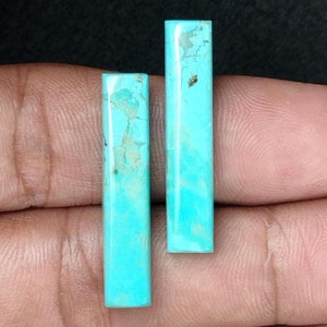 Könnte beinhalten: Zwei türkisfarbene Cabochons, geschnitten in rechteckige Formen, mit einer glatten, polierten Oberfläche. Die Steine sind hellblau mit braunen Adern und werden auf einer Hand gezeigt.