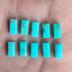 Może przedstawiać: Dziesięć turkusowych kaboszonów, każdy o prostokątnym kształcie, ułożonych jest w dwóch rzędach po pięć na dłoni. Kamienie mają żywy, niebiesko-zielony kolor.