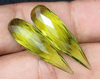 May include: Two faceted, elongated gemstones with a light yellow-green hue. The gems have a teardrop shape with a pointed end and a rounded, multi-faceted top. The gems are displayed against a neutral background.