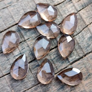 Pu&ograve; includere: Una collezione di 10 perle di pietra preziosa sfaccettate a forma di goccia di colore marrone chiaro. Le perle sono disposte su una superficie di legno.