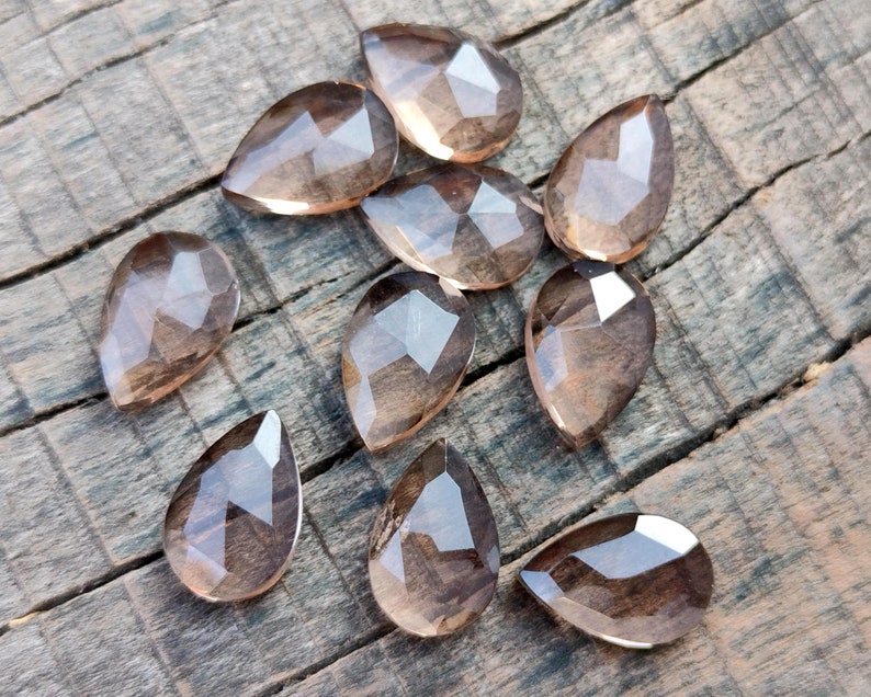 Pu&ograve; includere: Una collezione di 10 perle di pietra preziosa sfaccettate a forma di goccia di colore marrone chiaro. Le perle sono disposte su una superficie di legno.