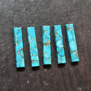 Può includere: Cinque pietre rettangolari turchesi con venature color oro. Le pietre sono di un blu brillante e sono disposte in fila. Le venature dorate creano un contrasto sorprendente con il blu. Queste pietre potrebbero essere utilizzate per la creazione di gioielli o altri lavori artigianali.