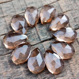 Pu&ograve; includere: Una collezione di dodici perle di pietra preziosa sfaccettate a forma di goccia di colore marrone chiaro. Le perle sono disposte in cerchio su una superficie di legno.