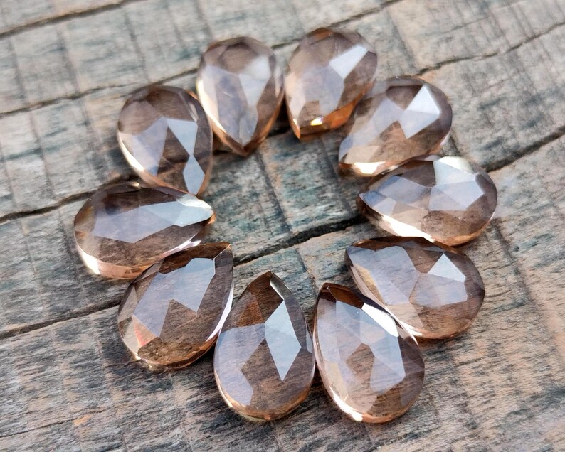 Pu&ograve; includere: Una collezione di dodici perle di pietra preziosa sfaccettate a forma di goccia di colore marrone chiaro. Le perle sono disposte in cerchio su una superficie di legno.