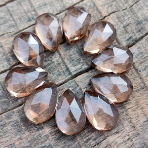 Pu&ograve; includere: Dieci cristalli di quarzo affumicato sfaccettati a forma di goccia in varie tonalit&agrave; di marrone. I cristalli sono disposti in un cerchio su una superficie di legno.