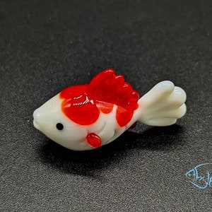 Peut inclure: Une petite figurine de poisson en verre blanc avec des marques rouges. Le poisson a un corps rond et une nageoire caudale.
