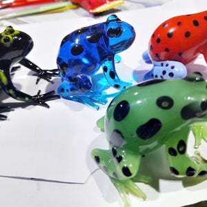 以下が含まれることがあります： カラフルなガラス製のカエルフィギュア4体。1つは黄色の斑点のある黒、もう1つは黒い斑点のある青、3つ目は黒い斑点のある赤、最後は黒い斑点のある緑です。各カエルは詳細な足を持っており、座った姿勢です。