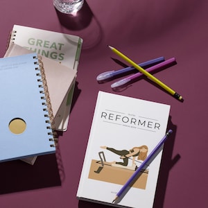 Puede incluir: Una pila de cuadernos, un vaso de agua y utensilios de escritura sobre una superficie púrpura. El cuaderno superior dice "GREAT THINGS". Es visible un manual de Pilates, con la palabra "REFORMER" en la portada.