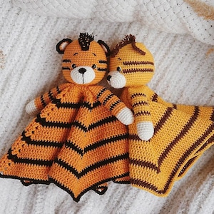 May include: Two crocheted tiger security blankets featuring orange and black striped bodies with round heads, small ears, and white muzzles. Each toy has a soft, textured yarn construction with embroidered facial features and small white paws.