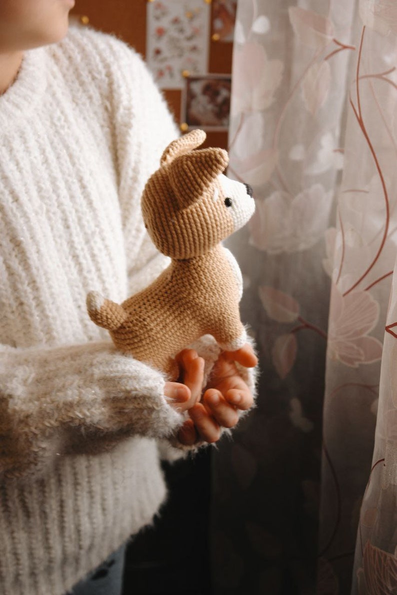 Peut inclure: Une personne tenant un jouet de chien en crochet marron et blanc. Le jouet a des yeux noirs et un nez brun.