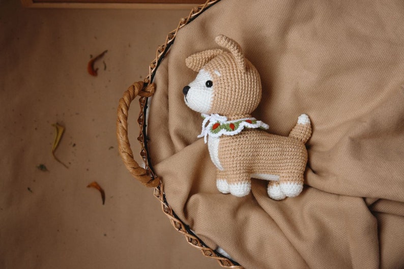 Peut inclure: Un jouet en peluche en forme de chien crochet&eacute; avec un collier blanc et vert. Le chien est brun clair et a un visage blanc. Le jouet est assis dans un panier en osier doubl&eacute; d'une couverture brune.