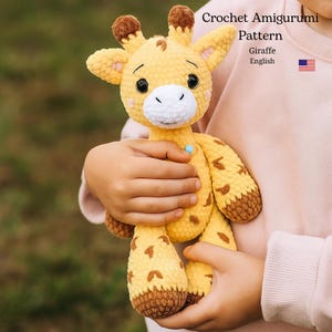 May include: A crocheted giraffe amigurumi in shades of yellow and brown is held in a person's hands. The giraffe has a white muzzle, black eyes, and brown spots. The text "Crochet Amigurumi Pattern" and "Giraffe English" is visible.
