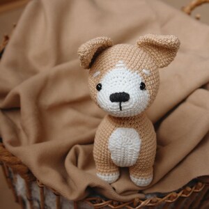 Peut inclure: Un jouet en peluche en forme de chien crochet&eacute; avec de la fourrure brune et blanche, assis dans un panier en osier doubl&eacute; d'un tissu brun.