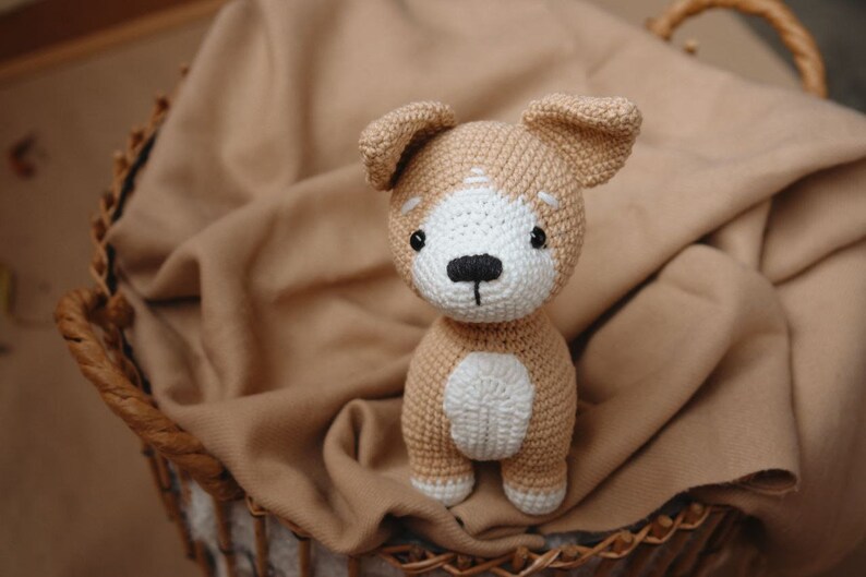 Peut inclure: Un jouet en peluche en forme de chien crochet&eacute; avec de la fourrure brune et blanche, assis dans un panier en osier doubl&eacute; d'un tissu brun.