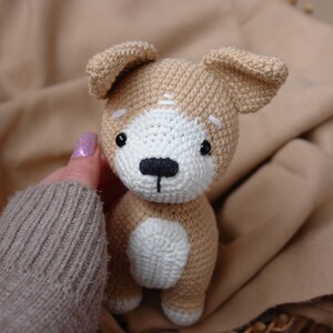 Peut inclure: Un jouet en peluche de chien crochet&eacute; avec de la fourrure brune et blanche, des yeux noirs et un nez noir. Le chien est assis sur une couverture brune.