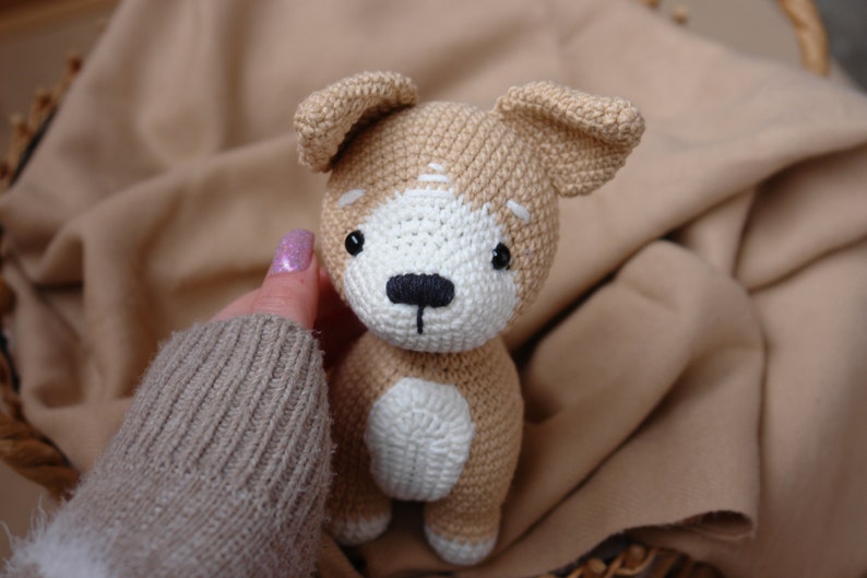 Peut inclure: Un jouet en peluche de chien crochet&eacute; avec de la fourrure brune et blanche, des yeux noirs et un nez noir. Le chien est assis sur une couverture brune.