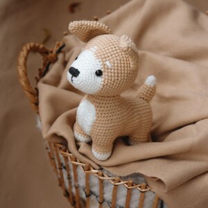 Peut inclure: Un jouet en peluche en crochet en forme de chien, de couleur marron et blanche, avec un nez noir. Le chien est assis dans un panier en osier doubl&eacute; d'une couverture marron.