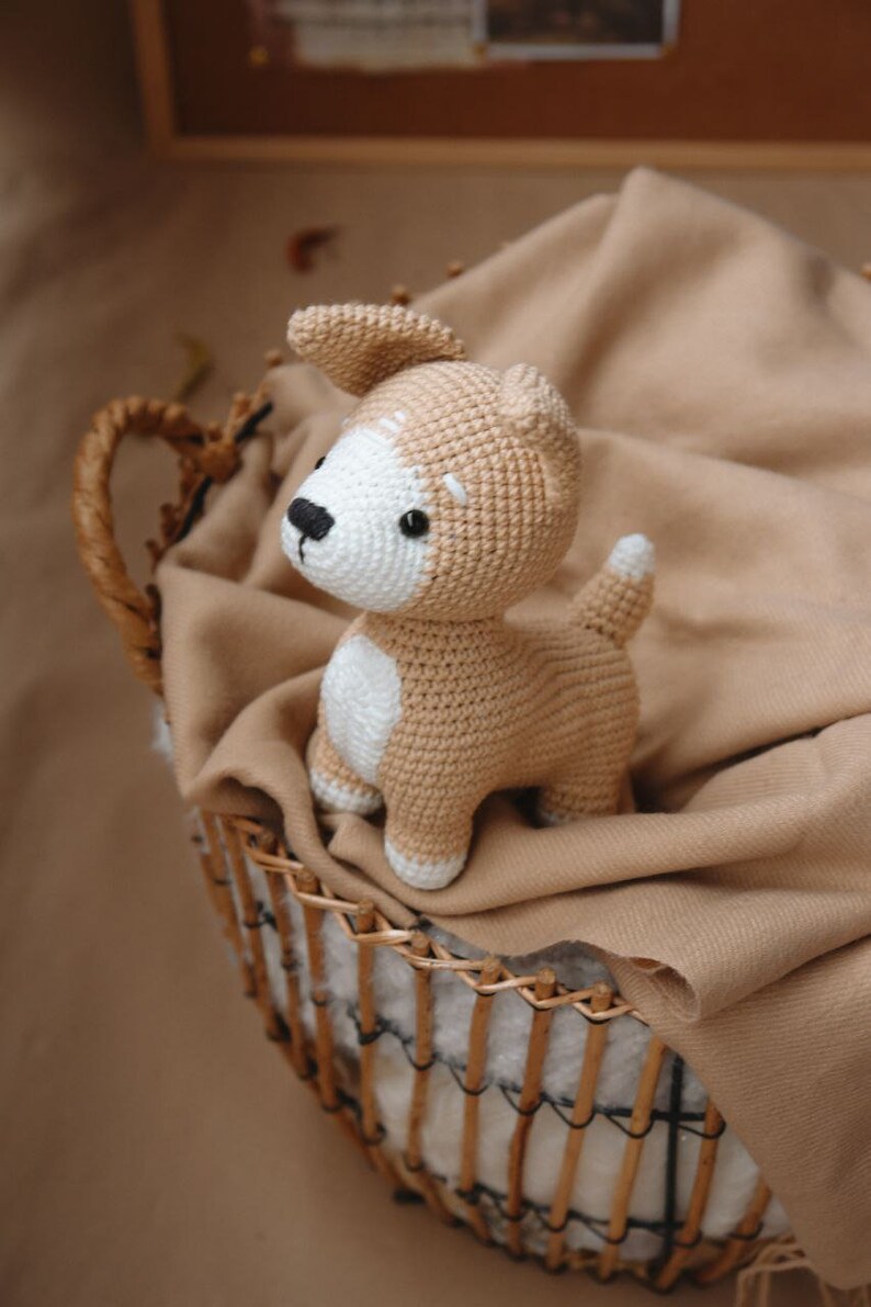 Peut inclure: Un jouet en peluche en crochet en forme de chien, de couleur marron et blanche, avec un nez noir. Le chien est assis dans un panier en osier doubl&eacute; d'une couverture marron.
