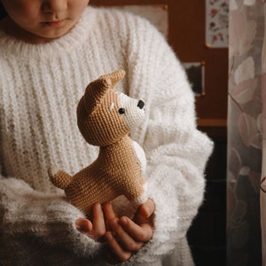 Peut inclure: Un enfant portant un pull blanc tient un jouet en crochet en forme de chien brun et blanc.