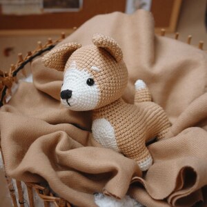 Peut inclure: Un jouet en peluche en crochet en forme de chien avec une fourrure brune et blanche. Le chien est assis sur une couverture brune.