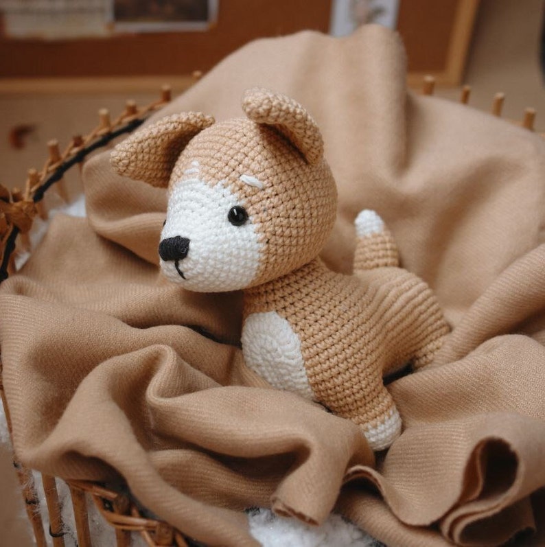 Peut inclure: Un jouet en peluche en crochet en forme de chien avec une fourrure brune et blanche. Le chien est assis sur une couverture brune.