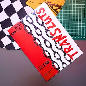 Puede incluir: Varios materiales impresos, incluyendo un folleto con la palabra "TRANS LUTS" en rojo, un gráfico de cadena y el texto "UN FINE TRANS-EROTICO". Un patrón de tablero de ajedrez en blanco y negro es visible en el fondo.
