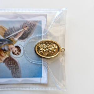 May include: A gold-coloured metal charm with the words "St. Michael" engraved on it. The charm is in a clear plastic bag with a white card featuring a religious image.