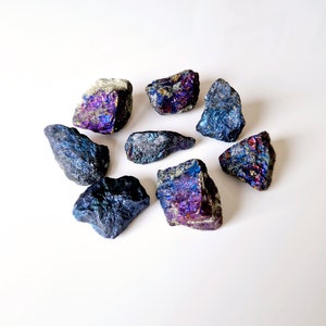 May include: A collection of eight iridescent pyrite stones, with shades of blue, purple, and gold, arranged in a circle on a white background.