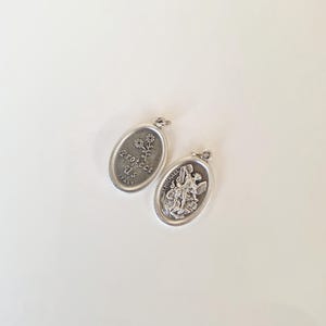 Op de afbeelding: Twee ovale zilveren hangers. Eén hanger heeft de tekst "Protect Us ITALY" met een bloemmotief. De andere hanger toont een afbeelding van St. Michael. Beide hangers hebben een kleine lus aan de bovenkant voor een ketting.