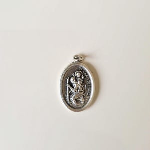 Könnte beinhalten: Ein ovaler Anhänger aus silberfarbenem Metall mit einem Relief, das den heiligen Christophorus zeigt, der ein Kind auf seinen Schultern trägt und einen Stab hält. Der Text ST. CHRISTOPHER ist auf der rechten Seite eingeprägt.