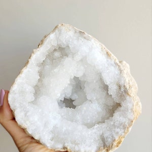 May include: A geode crystal with a beige, rough outer shell and a bright white, crystalline interior. The geode is open, displaying a cluster of crystals. The natural stone is held in a hand.