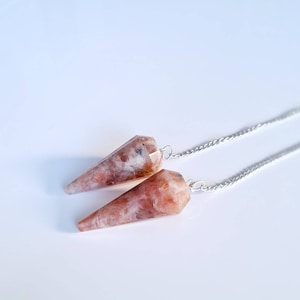 Può includere: Due pendoli di cristallo di pietra di sole con catene d'argento. I pendoli sono di colore rosa chiaro con macchie bianche e marroni.