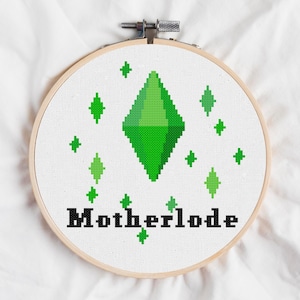May include: A cross-stitch embroidery hoop with a central green diamond and surrounding green diamond and star shapes. The word "Motherlode" is stitched in black below. The design is set on white fabric, held within a wooden embroidery hoop.