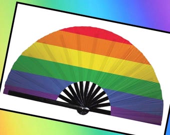 May include: A rainbow-colored hand fan with a black handle. The fan features the colors of the Pride flag: red, orange, yellow, green, blue, and purple. The fan is open, displaying its full arc.