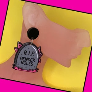 May include: A pair of earrings featuring a black tombstone with pink floral accents and the text "R.I.P. Gender Roles".