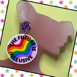 May include: A pair of earrings featuring a rainbow design and the text "The Future is Inclusive". The earrings are pink with a rainbow wave design and a purple glitter accent.