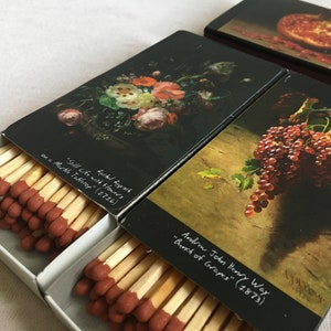 Pode incluir: Caixas de fósforos com estampas de arte. Uma apresenta uma natureza morta floral, outra um cacho de uvas e uma terceira uma romã. Os fósforos são visíveis em caixas abertas. O texto nas caixas de fósforos diz "Still Life with Flowers" e "Bunch of Grapes."