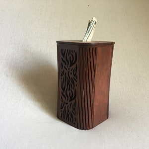 May include: A wooden money box with a decorative cutout design. The box is brown and has a slot on the top for inserting money. A dollar bill is visible in the slot.