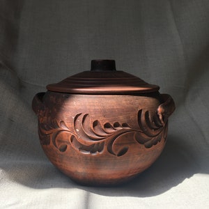 Può includere: Una pentola in ceramica marrone fatta a mano con coperchio, con disegni floreali intagliati. La pentola ha due manici ed è adatta per cucinare e servire. La ceramica rustica è esposta su uno sfondo neutro.