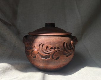 Könnte beinhalten: Ein handgefertigter, brauner Keramiktopf mit Deckel und geschnitzten floralen Mustern. Der Topf hat zwei Griffe und eignet sich zum Kochen und Servieren. Die rustikale Keramik wird auf neutralem Hintergrund präsentiert.