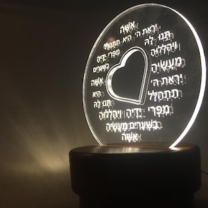 Puede incluir: Un disco de acrílico transparente con un recorte en forma de corazón, iluminado por una luz cálida. El disco presenta texto hebreo grabado alrededor del corazón y está montado sobre una base de madera oscura. Es una lámpara decorativa.