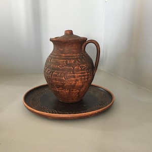 Può includere: Una brocca in ceramica marrone con coperchio e manico, appoggiata su un piatto abbinato. La brocca e il piatto presentano un design strutturato e intagliato. Il set è realizzato in terracotta ed è adatto per servire acqua o altre bevande.