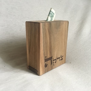 Peut inclure: Une boîte à monnaie en bois avec une fente pour les billets. La boîte est gravée du mot hébreu "Tzedakah" qui signifie charité.