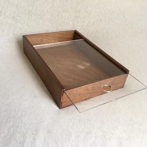 May include: A rectangular wooden box with a clear acrylic sliding lid. The box is made of a light brown wood and has a small, circular handle on the lid. The box is on a white surface.