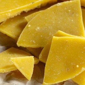 May include: Close-up of several pieces of yellow beeswax, broken into irregular shapes, on a brown paper surface.