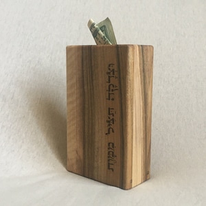 May include: A wooden box with a slot for money, engraved with Hebrew text. The box is made of light brown wood with a natural grain pattern.