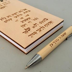 Può includere: Un taccuino in legno color chiaro con testo ebraico e una penna abbinata. La penna ha dettagli argentati e testo ebraico. Il taccuino è aperto, rivelando pagine bianche. La penna è accanto al taccuino.