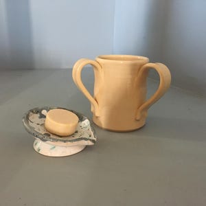 Puede incluir: Un jabonera de cerámica hecha a mano con una pastilla de jabón y una taza a juego. La jabonera es de color gris claro con detalles azules y blancos. La taza es de color melocotón claro y tiene dos asas.